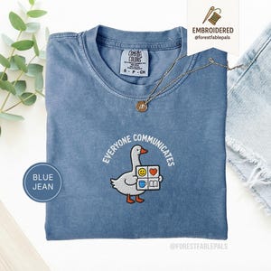 May include: Blue jean coloured t-shirt with the embroidered phrase "EVERYONE COMMUNICATES" around a cartoon goose holding a book and emoji blocks. The shirt has a crew neck and short sleeves. A gold necklace is draped across the shirt.