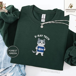 Könnte beinhalten: Dunkelgrünes Sweatshirt mit einem gestickten Design. Das Design zeigt eine grau-weiße Katze, die ein blau-weißes Röntgenbild hält, mit dem Text "X-RAY TECH" darüber. Eine silberne Halskette mit einem kleinen Anhänger ist ebenfalls sichtbar.