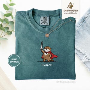 May include: Blue spruce t-shirt with an embroidered otter design. The otter is depicted holding a sword and wearing a red cape. The word "Huzzah" is embroidered below the otter. A gold necklace with a pendant is also visible.
