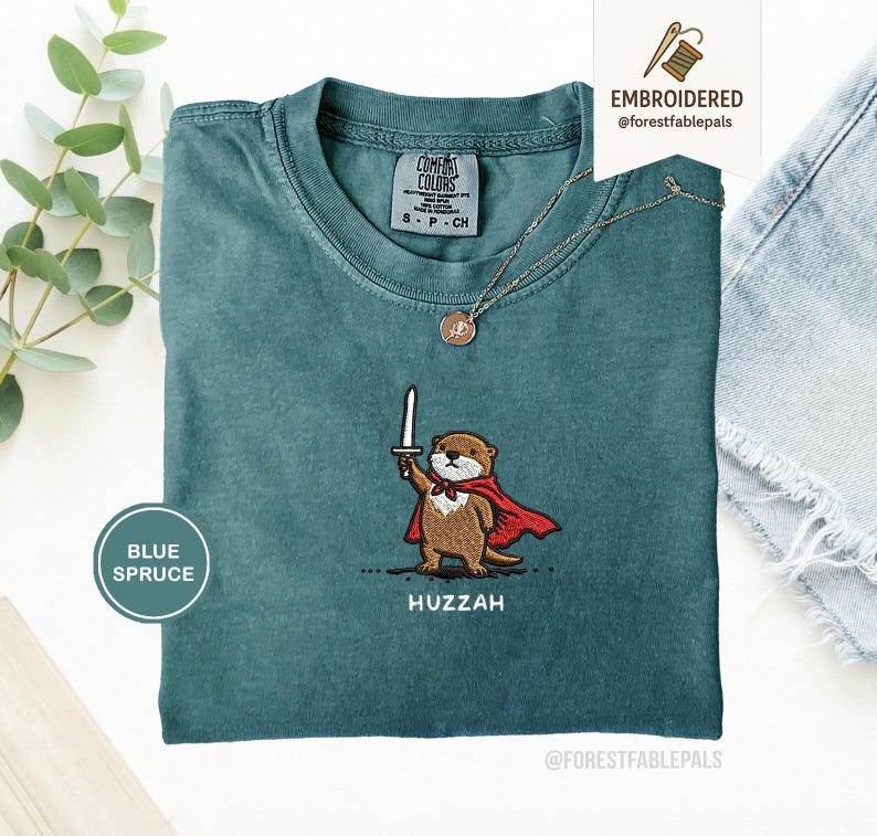 May include: Blue spruce t-shirt with an embroidered otter design. The otter is depicted holding a sword and wearing a red cape. The word "Huzzah" is embroidered below the otter. A gold necklace with a pendant is also visible.