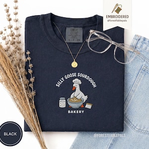 Peut inclure: T-shirt bleu marine avec un motif brodé d'une oie portant un chapeau de chef, mélangeant de la pâte, et le texte "Silly Goose Sourdough Bakery". Un collier doré et des lunettes transparentes sont posés sur le t-shirt. Le t-shirt est étiqueté "S, P, CH."