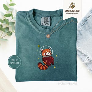 Op de afbeelding: Blauwgroen T-shirt met een geborduurde rode panda met een ruimtehelm. Het shirt heeft een ronde hals en korte mouwen. Het label zegt "COMFORT COLORS". Een gouden ketting met een hanger is ook zichtbaar.