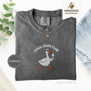 May include: A gray Comfort Colors t-shirt with the embroidered phrase "COOL DADS CLUB" and a cartoon goose wearing sunglasses and holding an ice cream cone. A gold necklace with a pendant is draped on the shirt.