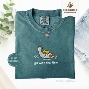 以下が含まれることがあります： 青いトウヒ色のTシャツで、オレンジ色のくちばしと足を持つ白いガチョウが背中に小さな黄色いアヒルを乗せている刺繍が施されています。刺繍の下には、白い文字で go with the flow と書かれています。