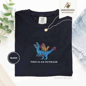 May include: Black t-shirt with an embroidered design of a brown cat riding a blue dinosaur with pink accents. The text "THIS IS AN OUTRAGE" is below the design. The shirt has a crew neck and a tag that says "COMFORT COLORS".