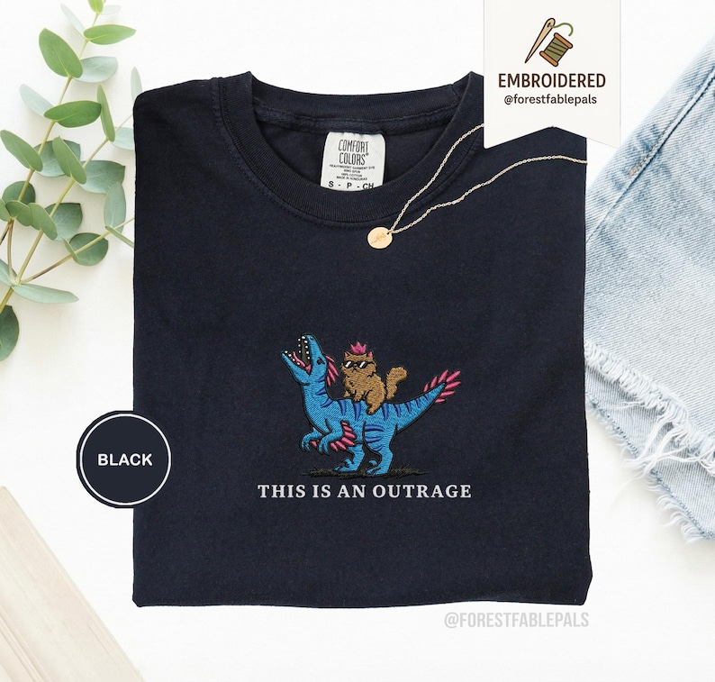 May include: Black t-shirt with an embroidered design of a brown cat riding a blue dinosaur with pink accents. The text "THIS IS AN OUTRAGE" is below the design. The shirt has a crew neck and a tag that says "COMFORT COLORS".
