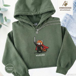 May include: Military green hoodie with a cartoon red panda holding a sword and wearing a red cape. The word "Huzzah" is printed below the panda. The hoodie has a hood and a silver chain necklace.