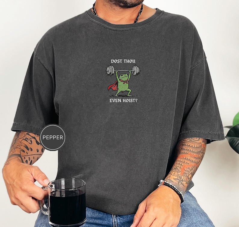 May include: Dark grey t-shirt with a cartoon frog lifting weights. The frog wears a red cape and bow tie, with the text "DOST THOU EVEN HOIST?" above and below the frog. The shirt has a Pepper brand label.