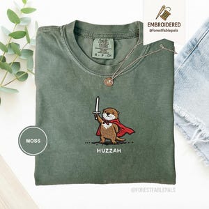 May include: Moss green t-shirt with an embroidered otter design. The otter is wearing a red cape and holding a sword, with the word "Huzzah" below. The shirt has a crew neck and short sleeves.