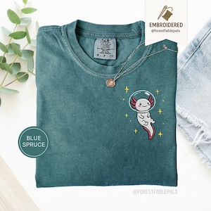 Könnte beinhalten: Blau-Fichte-T-Shirt mit einem gestickten Axolotl, das einen Weltraumhelm trägt. Das Shirt hat einen Rundhalsausschnitt und kurze Ärmel. Der Axolotl ist weiß mit rosa Kiemen und roter Kontur. Der Text "EMBROIDERED @forestfablepals" steht auf einem Etikett.