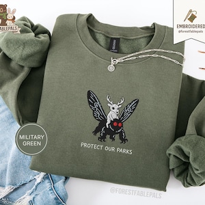 Peut inclure: Sweat-shirt vert militaire avec un motif brodé d'un cerf chevauchant un papillon de nuit, avec le texte "Protect Our Parks". Un collier argenté avec un petit pendentif est sur le sweat-shirt.