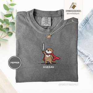 May include: A gray Comfort Colors t-shirt featuring an embroidered otter wearing a red cape and holding a sword. The word "Huzzah" is embroidered below the otter. A silver necklace with a pendant is on the shirt.