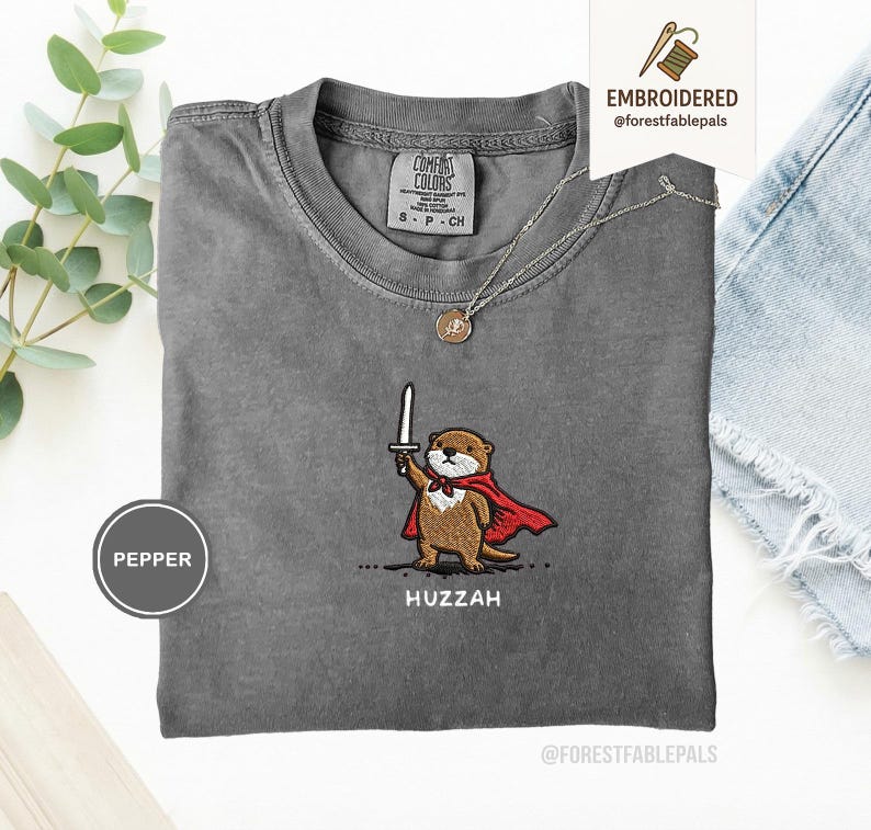 May include: A gray Comfort Colors t-shirt featuring an embroidered otter wearing a red cape and holding a sword. The word "Huzzah" is embroidered below the otter. A silver necklace with a pendant is on the shirt.