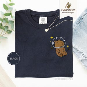 Peut inclure: T-shirt noir avec un motif brodé d'un capybara marron portant un casque spatial, entouré d'étoiles jaunes. Le t-shirt a un col rond et une petite étiquette. Un collier en or est drapé sur le t-shirt.