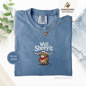 Könnte beinhalten: Ein blaues Kurzarm-T-Shirt mit einer Stickerei eines braunen Longhorn-Rinds, das einen weißen Cowboyhut und ein rotes Halstuch trägt. Der Text Well SHEEYIT ist über dem Rind gestickt. Das T-Shirt hat einen Rundhalsausschnitt und ein Etikett.