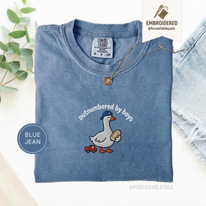 Peut inclure: T-shirt bleu jean avec un motif brodé d'une oie portant une casquette de baseball et tenant un ballon de football, avec le texte "outnumbered by boys". Une petite voiture rouge est également brodée. Le t-shirt a un col rond et des manches courtes.