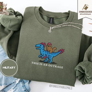 Puede incluir: Sudadera verde oliva con un diseño bordado de un dinosaurio azul con detalles rosas y un gato marrón con corona. El texto "THIS IS AN OUTRAGE" está debajo. Una cadena de plata está sobre la sudadera.