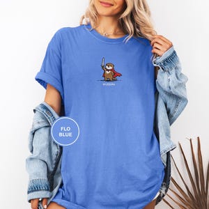 May include: Flo blue t-shirt featuring a cartoon otter with a sword and cape. The otter is wearing a red cape and holding a silver sword. The word "Huzzah" is printed below the otter. The shirt is a solid flo blue color.
