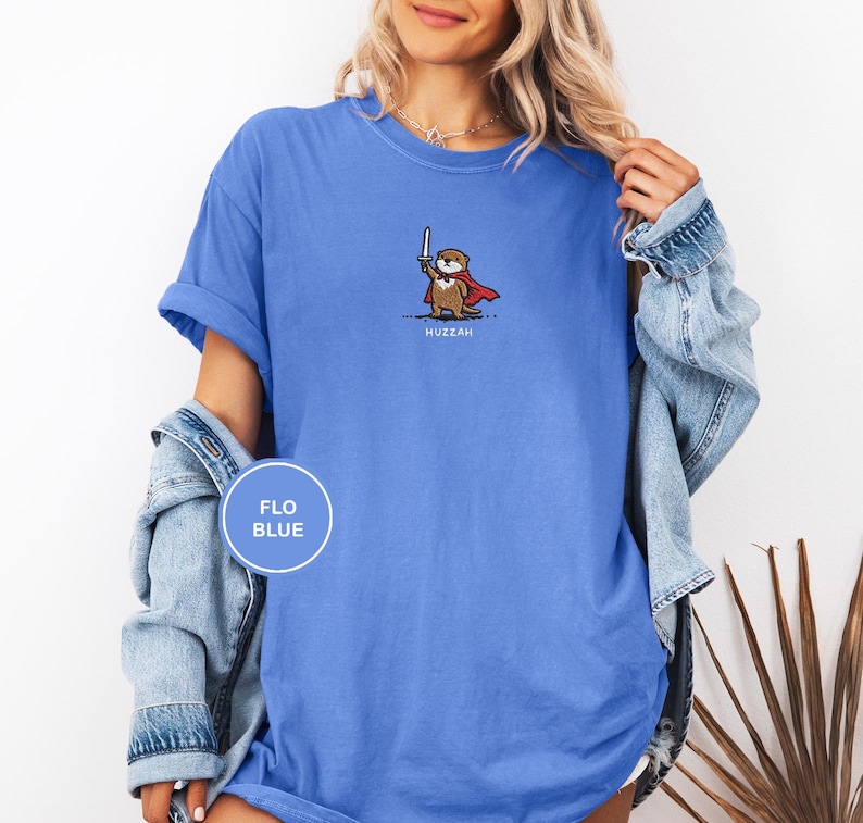 May include: Flo blue t-shirt featuring a cartoon otter with a sword and cape. The otter is wearing a red cape and holding a silver sword. The word "Huzzah" is printed below the otter. The shirt is a solid flo blue color.