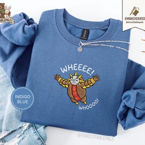 Könnte beinhalten: Indigoblaues Sweatshirt mit einem gestickten Cartoon-Charakter mit ausgestreckten Armen, dem Text "WHEEEE!" darüber und "WHOOOO!" darunter. Eine silberne Halskette mit einem runden Anhänger ist über dem Sweatshirt drapiert. Das Markenlogo befindet sich oben rechts.