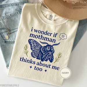 May include: An ivory t-shirt featuring a blue graphic of a mothman with the text "i wonder if mothman thinks about me too" and a small thought bubble with a heart. The design includes two small green sprigs and two four-pointed stars.