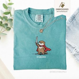 May include: A teal Comfort Colors t-shirt featuring an embroidered otter wearing a red cape and holding a sword. The word "Huzzah" is embroidered below the otter. A gold necklace with a pendant is draped on the shirt.