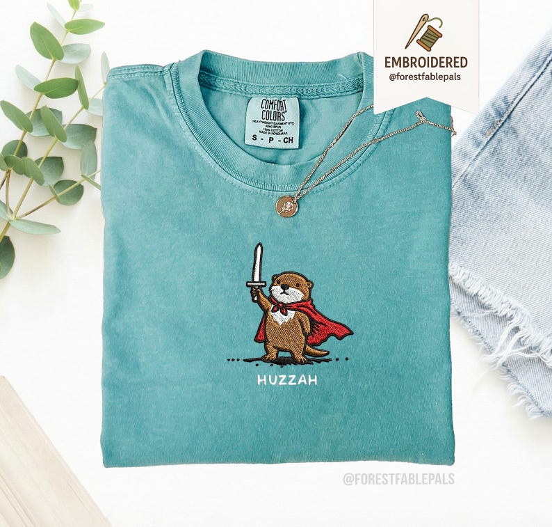 May include: A teal Comfort Colors t-shirt featuring an embroidered otter wearing a red cape and holding a sword. The word "Huzzah" is embroidered below the otter. A gold necklace with a pendant is draped on the shirt.