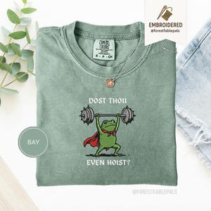 May include: A sage green t-shirt featuring a cartoon frog lifting weights, with a cape and bow tie. The text reads "DOST THOU EVEN HOIST?" The shirt has a crew neck and short sleeves. A silver necklace with a pendant is visible.