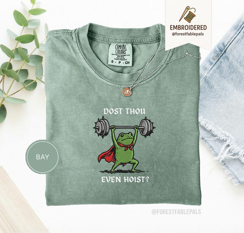 May include: A sage green t-shirt featuring a cartoon frog lifting weights, with a cape and bow tie. The text reads "DOST THOU EVEN HOIST?" The shirt has a crew neck and short sleeves. A silver necklace with a pendant is visible.