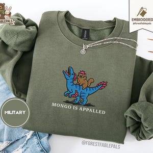 May include: Olive green sweatshirt with an embroidered design featuring a blue dinosaur with a brown cat wearing a crown. The text "MONGO IS APPALLED" is below the design. A silver necklace is draped across the sweatshirt.