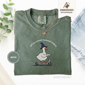 Op de afbeelding: Mosgroen T-shirt met de geborduurde tekst "Off to commit husband foolery". Het ontwerp toont een cartoon eend met een heksenhoed op een scooter. Het shirt heeft een ronde hals en korte mouwen.