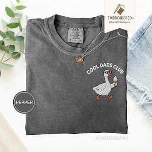 May include: A charcoal gray t-shirt with the embroidered phrase "COOL DADS CLUB" and a cartoon goose wearing sunglasses and holding an ice cream cone. The shirt has a crew neck and a small tag on the upper back.