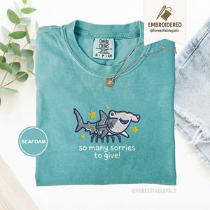 Może przedstawiać: Miętowa koszulka z haftowanym wzorem rekina młota i napisem "so many sorries to give!". Koszulka ma okrągły dekolt i krótkie rękawy. Złoty naszyjnik z wisiorkiem jest zarzucony na koszulkę.