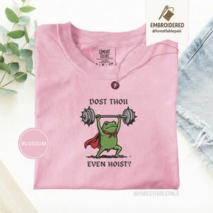 May include: A pale pink t-shirt featuring the embroidered phrase "DOST THOU EVEN HOIST?" and a green frog lifting weights. The frog wears a red cape and bow tie. The shirt has a crew neck and short sleeves.