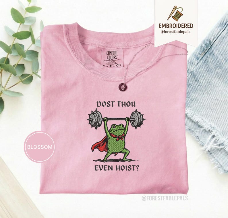 May include: A pale pink t-shirt featuring the embroidered phrase "DOST THOU EVEN HOIST?" and a green frog lifting weights. The frog wears a red cape and bow tie. The shirt has a crew neck and short sleeves.