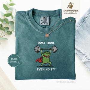 May include: Blue spruce t-shirt featuring a frog lifting weights, wearing a red cape and green bow tie. The text on the shirt reads "DOST THOU EVEN HOIST?" and "EMBROIDERED @forestfablepals".