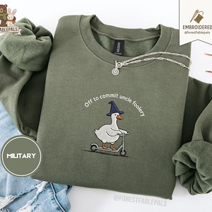 May include: An olive green sweatshirt featuring an embroidered goose wearing a witch's hat and riding a scooter. The text reads "Off to commit uncle foolery". A silver necklace is draped across the sweatshirt.