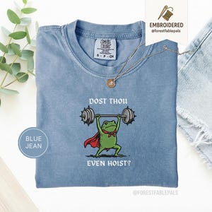 May include: A blue jean Comfort Colors t-shirt featuring a frog lifting weights. The frog is depicted with a red cape and a green bow tie. The text on the shirt reads "DOST THOU EVEN HOIST?" A gold necklace is draped on the shirt.