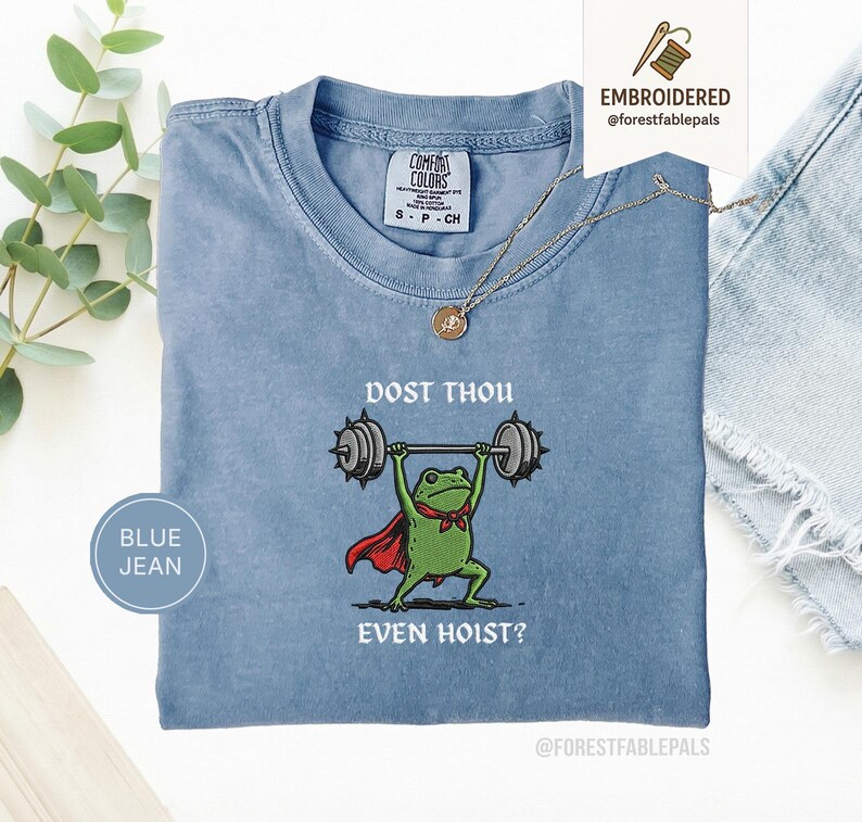 May include: A blue jean Comfort Colors t-shirt featuring a frog lifting weights. The frog is depicted with a red cape and a green bow tie. The text on the shirt reads "DOST THOU EVEN HOIST?" A gold necklace is draped on the shirt.