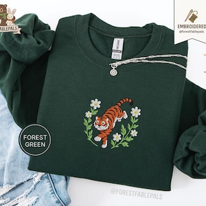 Pode incluir: Camisola verde floresta com um desenho de tigre bordado rodeado por flores brancas e folhas verdes. O tigre é laranja e preto. Um colar de prata com um pingente de sol está na camisola. O texto "FOREST GREEN" está na camisola.