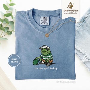 Puede incluir: Una camiseta azul claro con un diseño de rana bordada. La rana lleva una camisa a rayas verdes y blancas y una visera blanca, sosteniendo un disco verde. El texto en la camiseta dice "no disc golf today."