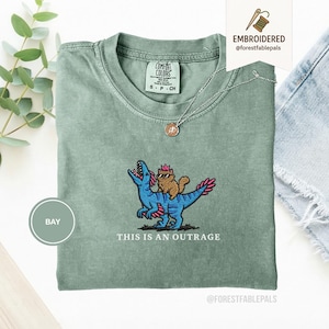 May include: A sage green t-shirt with an embroidered design of a brown cat wearing sunglasses riding a blue dinosaur. The text "THIS IS AN OUTRAGE" is below the design. The shirt has a crew neck and short sleeves.