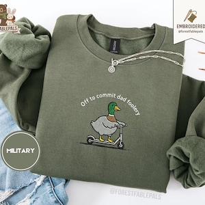 Op de afbeelding: Olijfgroene sweatshirt met de geborduurde tekst "Off to commit dad foolery" en een cartoon eend op een scooter. Een zilveren ketting met een kleine hanger is over het sweatshirt gedrapeerd. Het sweatshirt heeft een militaire groene cirkelgrafiek.