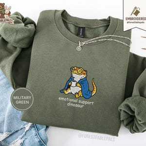 Könnte beinhalten: Militärgrünes Sweatshirt mit einem gestickten Design einer Eidechse mit Brille und blauem Umhang, die eine Tasse hält. Der Text "emotional support dinosaur" steht unter der Eidechse. Eine silberne Halskette liegt auf dem Sweatshirt.