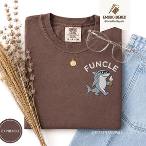 May include: A brown t-shirt with the word "FUNCLE" embroidered in white above a cartoon shark wearing sunglasses and holding an ice cream cone. The shirt has a crew neck and short sleeves. A gold necklace and clear glasses are also visible.