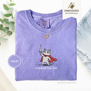Puede incluir: Camiseta violeta Comfort Colors con un diseño bordado de un gato con capa roja sosteniendo una espada. El texto "CONQUER THE TEST" está debajo. Se ve un collar de oro.
