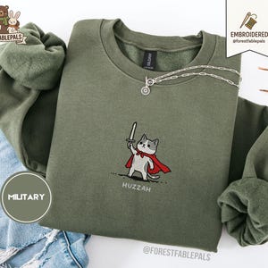 Puede incluir: Sudadera verde oliva con un gráfico de gato bordado en la parte delantera. El gato lleva una capa roja y sostiene una espada. La palabra "Huzzah" está bordada debajo. Un collar plateado está sobre la sudadera.
