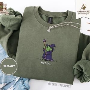 Puede incluir: Sudadera verde oliva con un bordado de una rana de dibujos animados vestida de mago, sosteniendo un bastón. La palabra "Huzzah" está bordada debajo de la rana. Un collar plateado con un colgante de sol está sobre la sudadera.