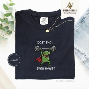 May include: Black crew neck t-shirt featuring a cartoon frog lifting weights. The frog is wearing a red cape and a green bow tie. The text on the shirt reads "DOST THOU EVEN HOIST?" A gold necklace and a "BLACK" circle are also visible.