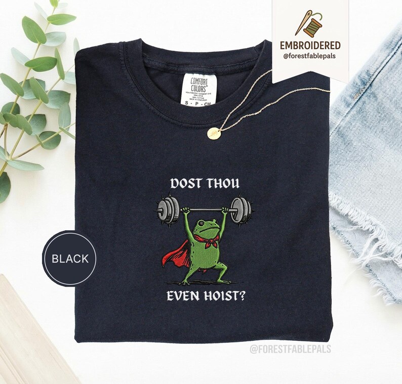 May include: Black crew neck t-shirt featuring a cartoon frog lifting weights. The frog is wearing a red cape and a green bow tie. The text on the shirt reads "DOST THOU EVEN HOIST?" A gold necklace and a "BLACK" circle are also visible.